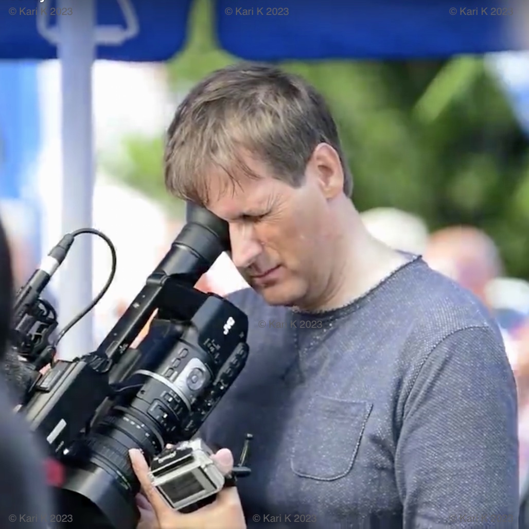 Valokuva henkilöstä videokuvaamassa ulkoilmatapahtumaa aurinkoisena kesäpäivänä.